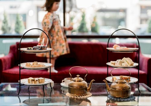 A cozy tea-time setup with a three-tier snack stand and two ornate teapots on a glass table, a blurred person in the background.