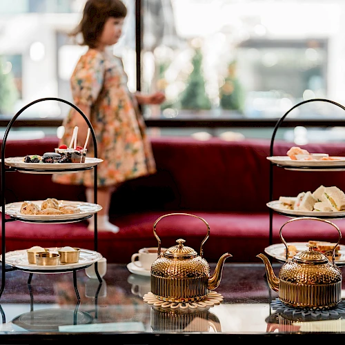 A cozy tea-time setup with a three-tier snack stand and two ornate teapots on a glass table, a blurred person in the background.