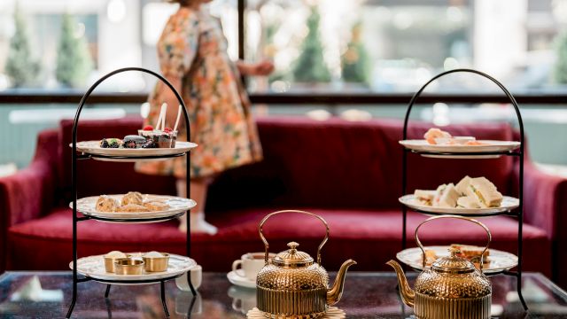 A cozy tea-time setup with a three-tier snack stand and two ornate teapots on a glass table, a blurred person in the background.