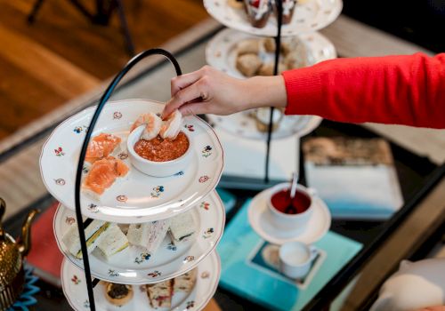 A hand in red selects bites from a tiered afternoon tea stand with pastries and cookies, alongside a scone and a cup of tea on the side.