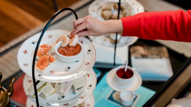 A hand in red selects bites from a tiered afternoon tea stand with pastries and cookies, alongside a scone and a cup of tea on the side.