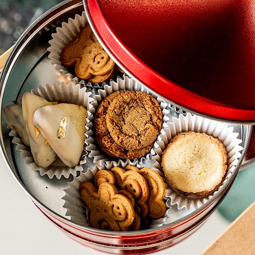 A red tin contains assorted cookies, including a heart-shaped one and animal-shaped ones, arranged in paper cups.