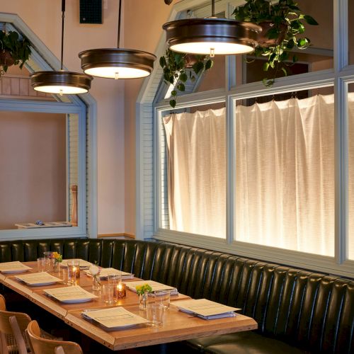 A cozy restaurant setting with a wooden table, chairs, booth seating, hanging lights, and windows framed with plants.