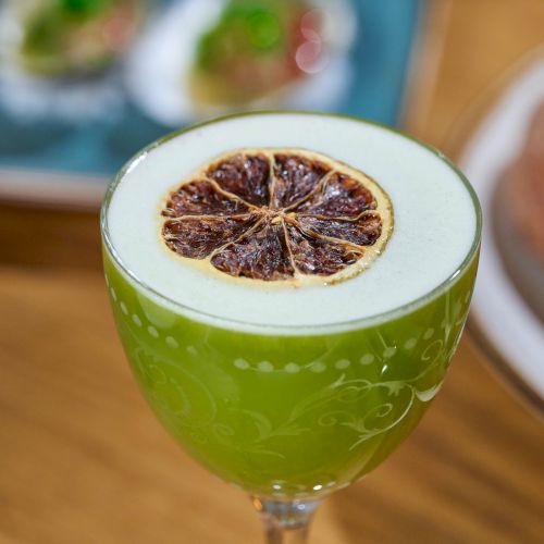 A green cocktail with a foamy top, garnished with a dried orange slice, sits in focus. Blurred background shows a plate and snacks.