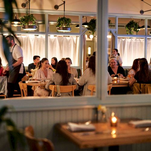 The image shows a busy restaurant with diners at tables, soft lighting, and a waiter serving. A candlelit table is in the foreground.