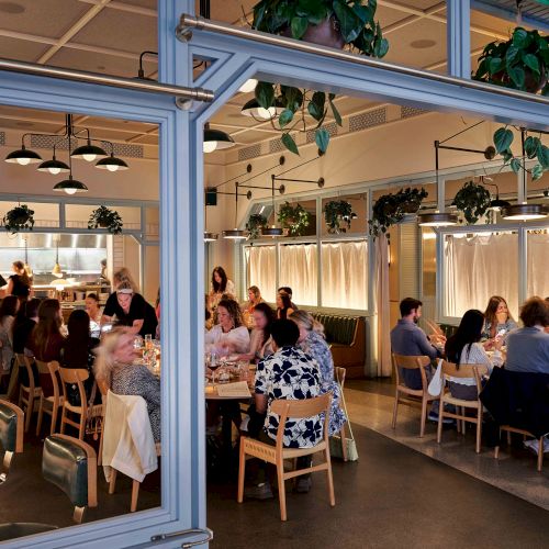 The image shows a lively restaurant with people dining at tables, surrounded by modern decor and hanging plants.
