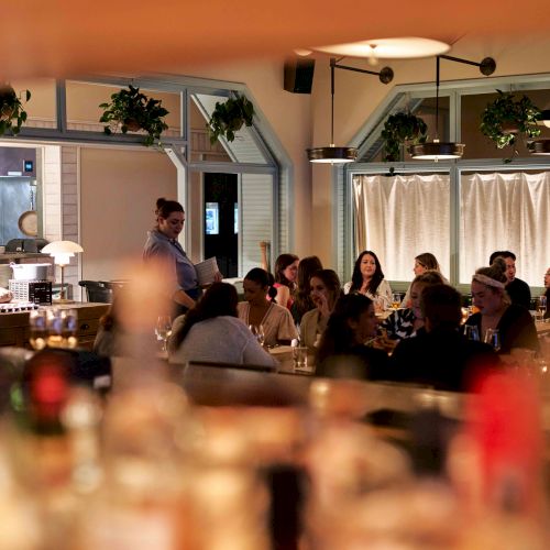 A cozy, bustling restaurant scene with patrons dining and conversing, featuring dim lighting and greenery decor for a warm atmosphere.