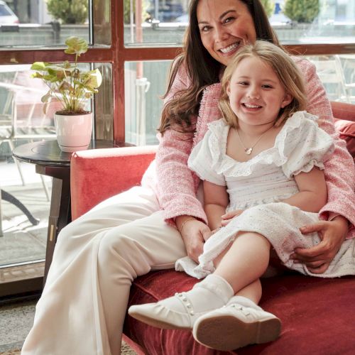 A woman and a child are smiling while sitting on a red couch in a room with large windows and a potted plant.