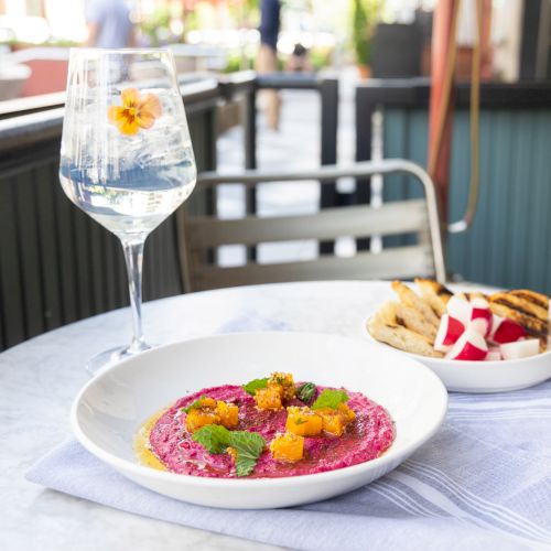 A table with beet hummus garnished with herbs and peaches, a side of toast with spread, and a glass of sparkling water with a flower.