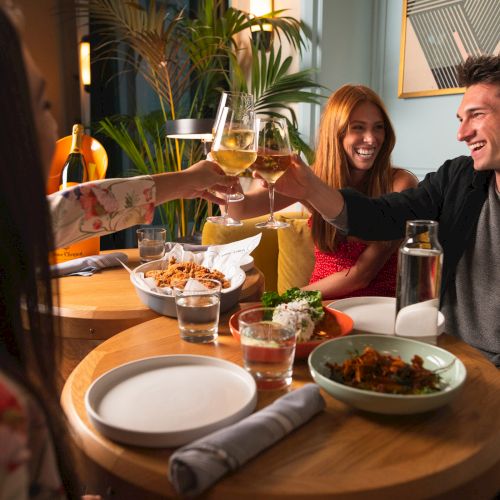 People are sitting at a table enjoying a meal, clinking glasses and laughing in a warmly lit restaurant or dining area.