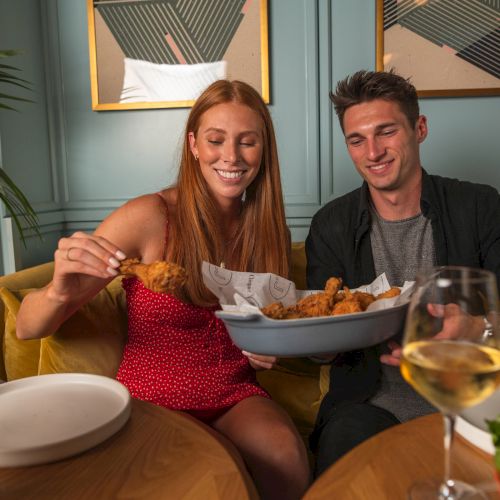 A woman and a man are sharing a plate of fried food at a dining table with glasses of white wine.