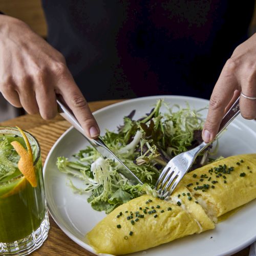 A person is cutting into an omelette with a side of greens, accompanied by a green beverage on a wooden table.