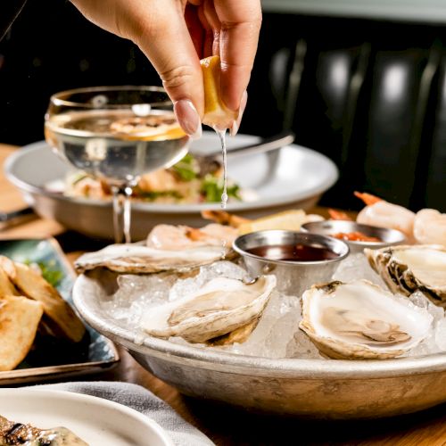 A hand squeezes lemon over a plate of oysters on ice; a glass of wine and other dishes are visible in the background.