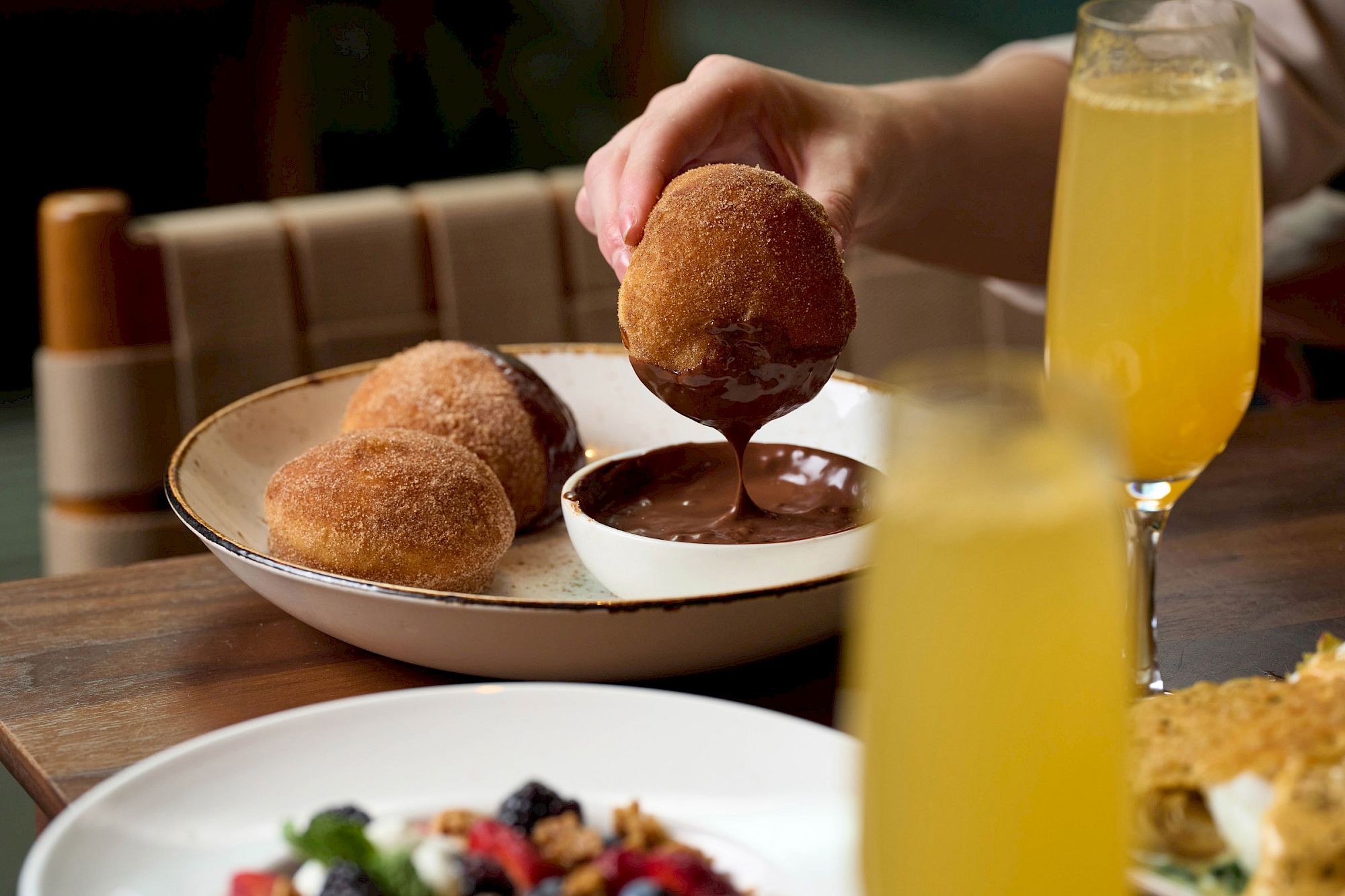 A person dips a doughnut-like pastry into chocolate sauce, surrounded by drinks and food on a table.