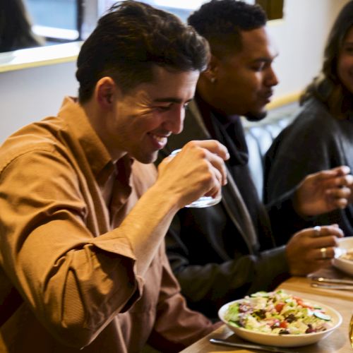 A person is enjoying food at a restaurant, holding a drink, with a salad bowl on the table, sitting next to others.