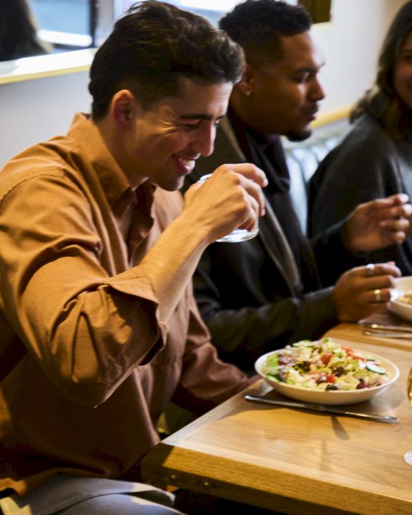 A person is enjoying food at a restaurant, holding a drink, with a salad bowl on the table, sitting next to others.