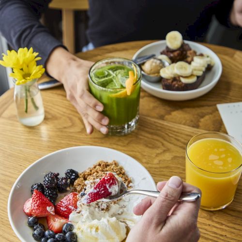 A breakfast setting with yogurt, berries, granola, green juice, orange juice, and a plate of bananas and toast, on a wooden table.