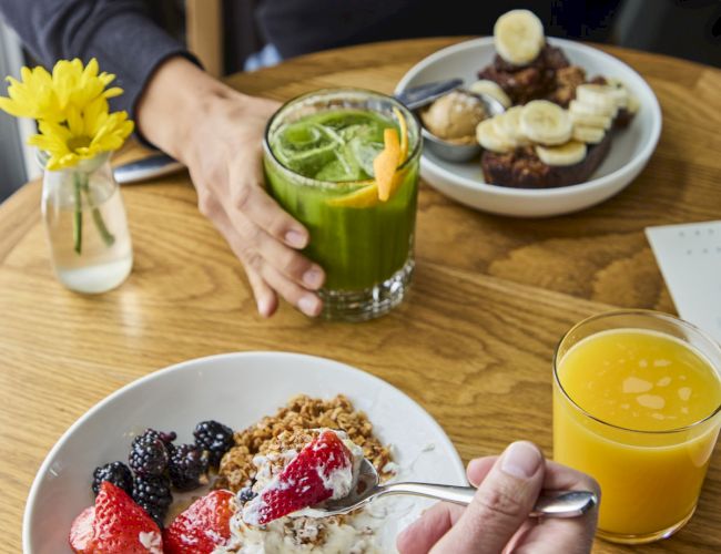 A breakfast setting with yogurt, berries, granola, green juice, orange juice, and a plate of bananas and toast, on a wooden table.