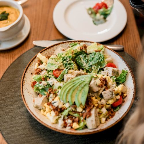 A colorful salad bowl with avocado slices on top, plus chopped veggies and eggs, served on a rustic plate with other dishes nearby.