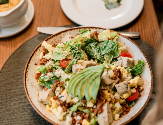 A colorful salad bowl with avocado slices on top, plus chopped veggies and eggs, served on a rustic plate with other dishes nearby.