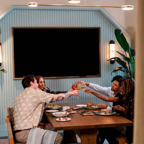 A group of friends clink glasses at a wooden dining table, sharing a meal in a cozy, plant-filled room with a blank wall TV behind them.