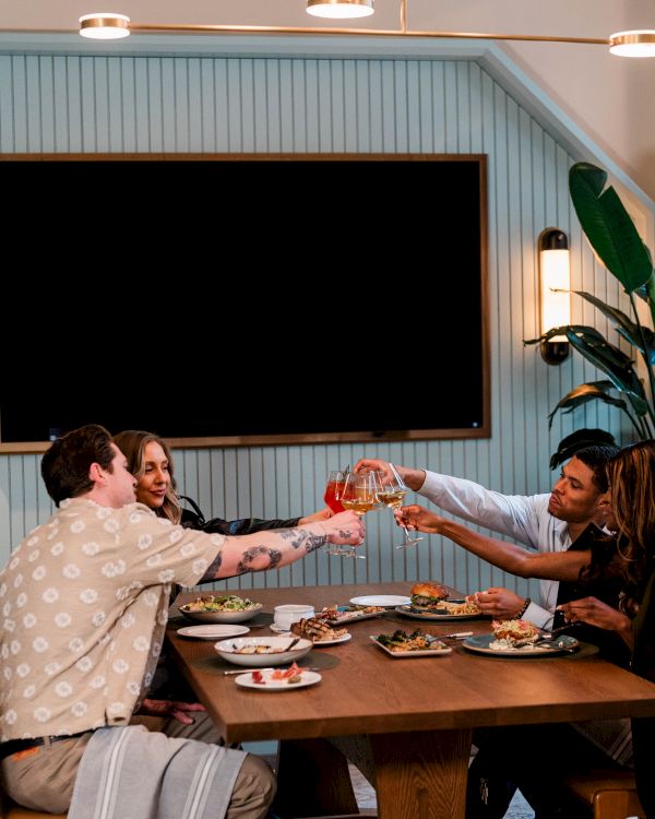A group of friends clink glasses at a wooden dining table, sharing a meal in a cozy, plant-filled room with a blank wall TV behind them.