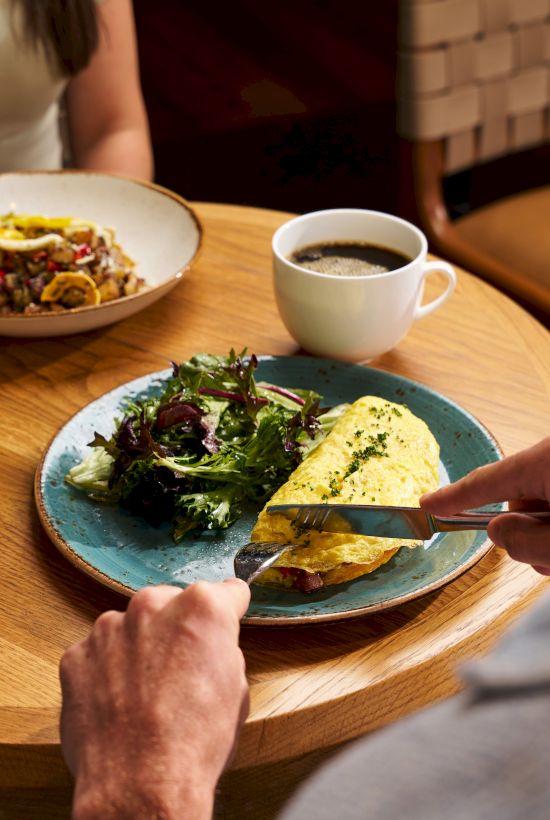 A person cutting into a herb-topped omelet or quiche on a blue plate, with a side salad, coffee cup, and another dish in the background at a round wooden table.