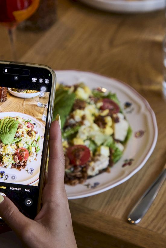 A person takes a photo of a salad with a smartphone at a wooden table set with glasses and utensils.