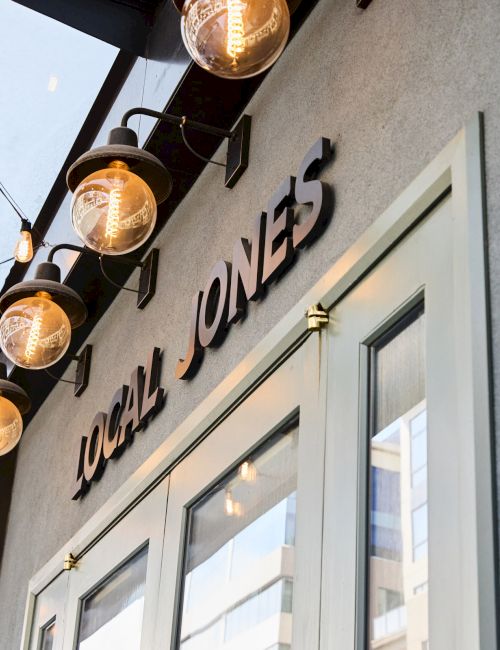 An exterior view of a building with the sign "LOCAL JONES" is shown, featuring decorative lighting and a partial glimpse of the entrance.