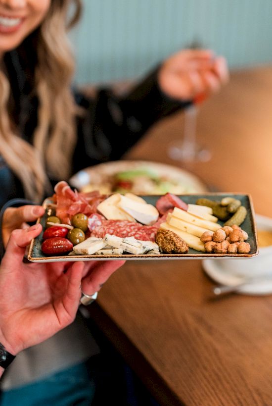 Two people share a charcuterie board with meats, cheeses, crackers, and olives, while chatting at a wooden table.