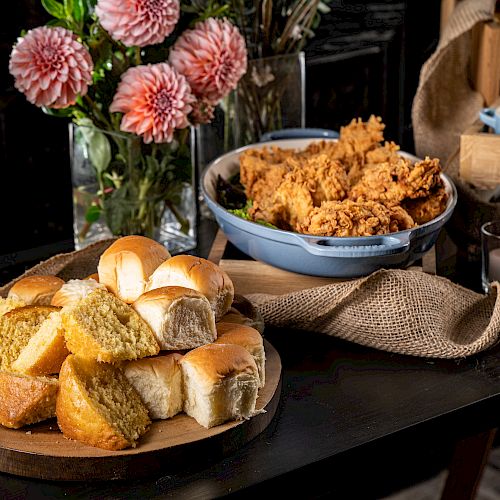 A table with bread rolls, fried chicken, flowers, and drinks, set on a dark surface with a rustic cloth.