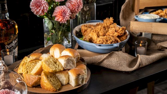 A table with bread rolls, fried chicken, flowers, and drinks, set on a dark surface with a rustic cloth.