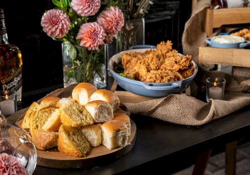 The image shows a table with bread, fried food in dishes, bottles, and pink flowers for decoration, arranged on a burlap cloth.