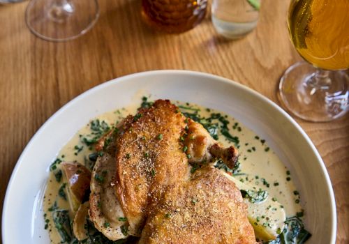 Crispy roasted chicken atop a bed of creamy herb sauce with mushrooms and greens, served on a white plate alongside wine and beer glasses.
