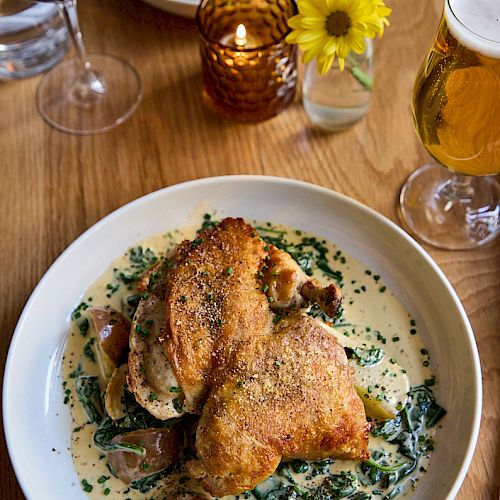 Crispy roasted chicken atop a bed of creamy herb sauce with mushrooms and greens, served on a white plate alongside wine and beer glasses.