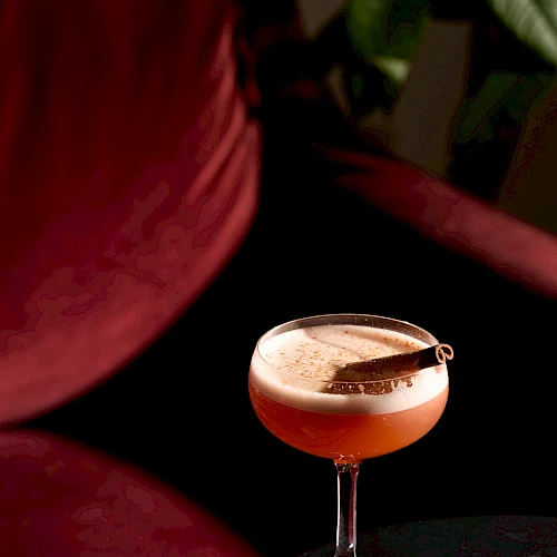 A cocktail in a coupe glass with a peachy-pink hue rests on a dark table, a red sofa and green leaves in the background, cozy vibe.