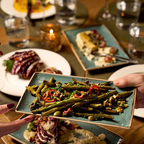 People sharing a colorful plated meal at a table, with a hand presenting a rectangular plate of saut&eacute;ed vegetables and greens, others nearby, drinks and more dishes on the table.