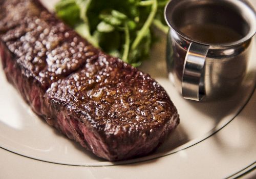 A seared steak with a brown crust on a plate, with a small cup of sauce on the side and some greens in the background.