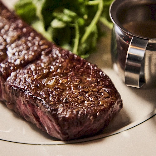 A seared steak with a brown crust on a plate, with a small cup of sauce on the side and some greens in the background.