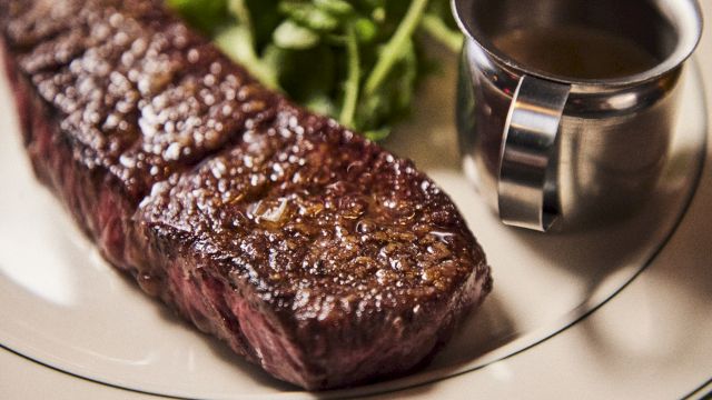 A seared steak with a brown crust on a plate, with a small cup of sauce on the side and some greens in the background.