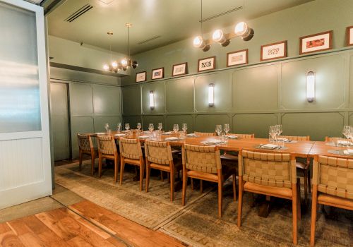 A stylish dining room with a long wooden table, cushioned chairs, warm pendant lights, and framed artwork along olive-green paneled walls.