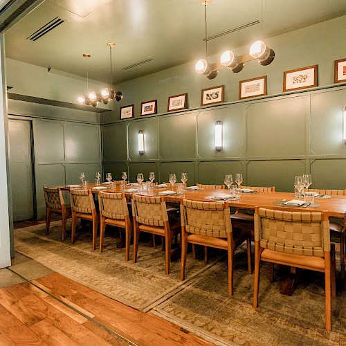A stylish dining room with a long wooden table, cushioned chairs, warm pendant lights, and framed artwork along olive-green paneled walls.