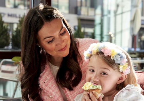 A woman and a little girl share a sweet moment, the girl wearing a flower headband and a white dress while biting a pastry.