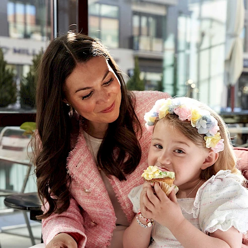 A woman and a little girl share a sweet moment, the girl wearing a flower headband and a white dress while biting a pastry.
