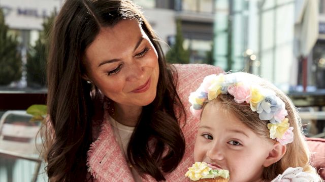 A woman and a little girl share a sweet moment, the girl wearing a flower headband and a white dress while biting a pastry.