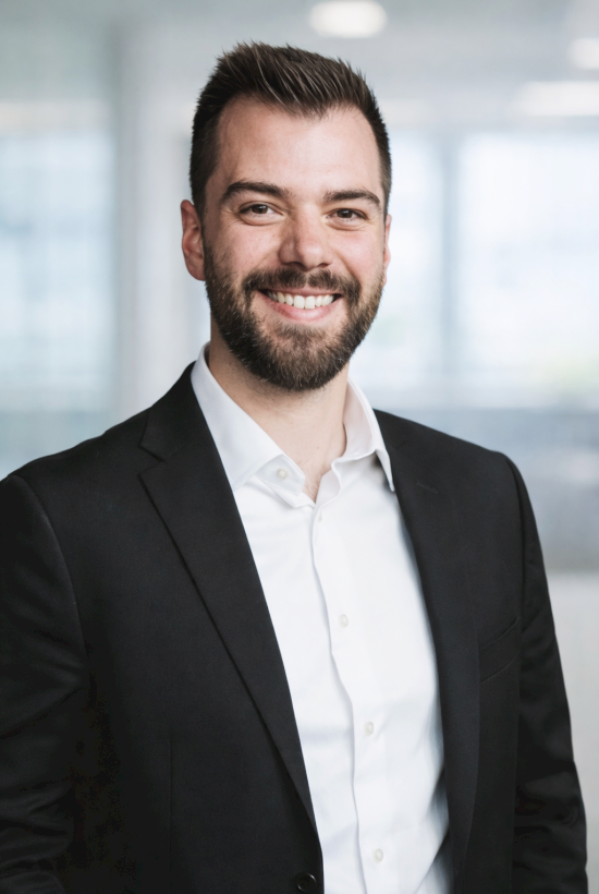 A confident businessman in a black suit and white shirt, smiling in a bright office environment.