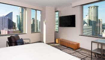 The image shows a modern hotel room with large windows offering a cityscape view, a bed, a TV on the wall, and contemporary furniture.