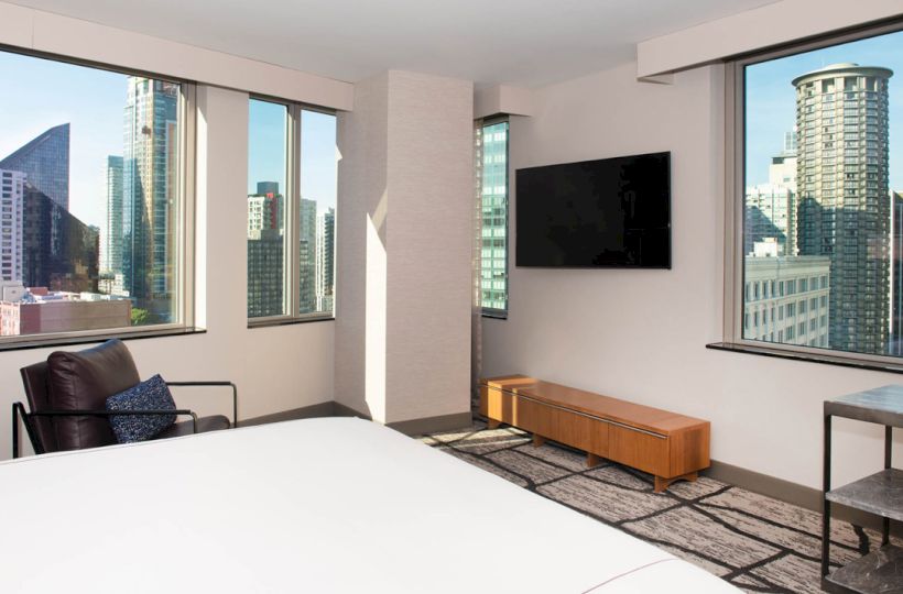The image shows a modern hotel room with large windows offering a cityscape view, a bed, a TV on the wall, and contemporary furniture.