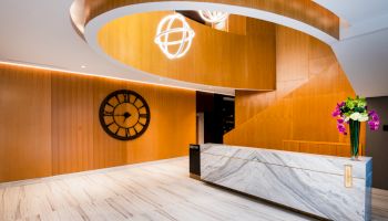 A modern lobby with a marble reception desk, wooden spiral staircase, spherical light fixture, wall clock, and a vase of colorful flowers.