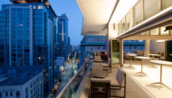 A modern rooftop terrace with seating overlooks a city street lined with tall buildings during twilight.